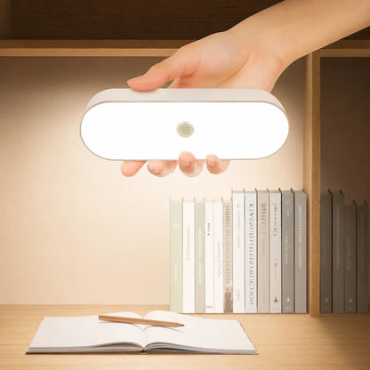 A hand holding a glowing white rechargeable motion sensor LED wall light, illuminating books and an open notebook on a wooden shelf.