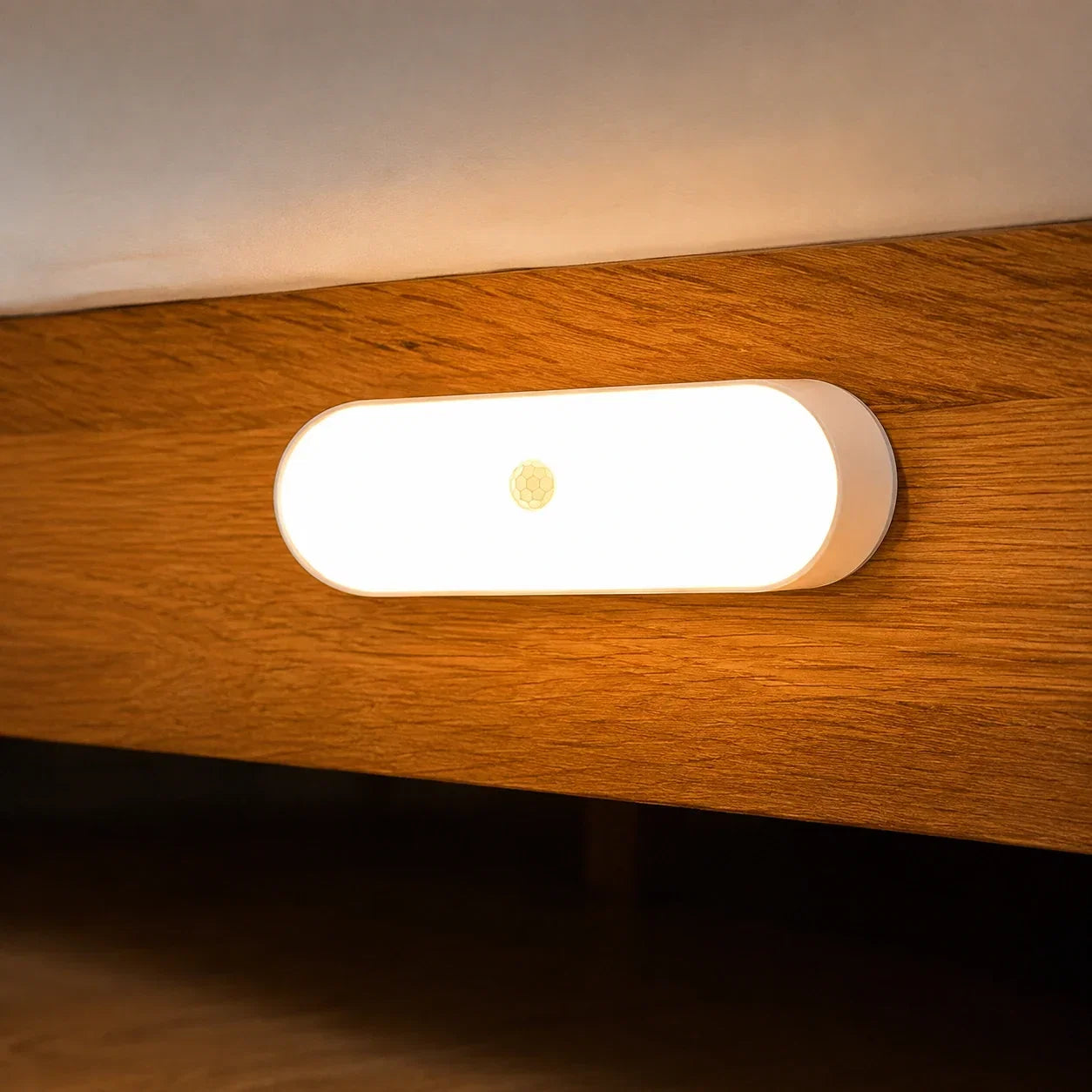 A sleek, oval rechargeable LED wall light with a motion sensor, emitting a warm glow while mounted on a wooden bed frame in a bedroom setting.
