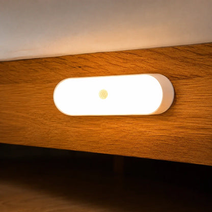 A sleek, oval rechargeable LED wall light with a motion sensor, emitting a warm glow while mounted on a wooden bed frame in a bedroom setting.