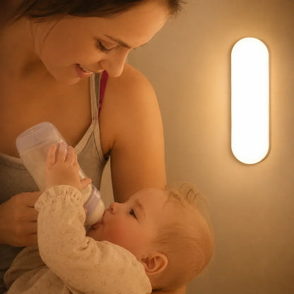 A mother bottle-feeding her baby in a nursery, illuminated by the warm glow of a rechargeable oval LED wall light mounted on the wall.