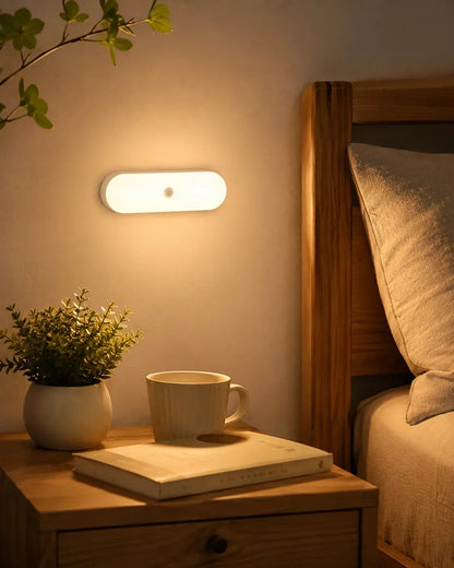 A rechargeable motion sensor LED wall light emitting a warm glow above a bedside table with a book, a mug, and a small potted plant in a bedroom.