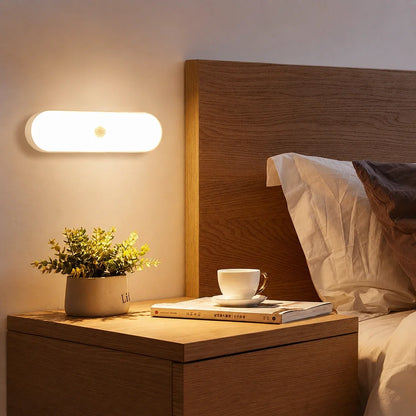 Rechargeable motion sensor LED wall light casting a warm glow above a wooden bedside table with a potted plant, book and teacup in a bedroom.