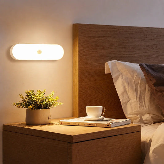Rechargeable motion sensor LED wall light casting a warm glow above a wooden bedside table with a potted plant, book and teacup in a bedroom.