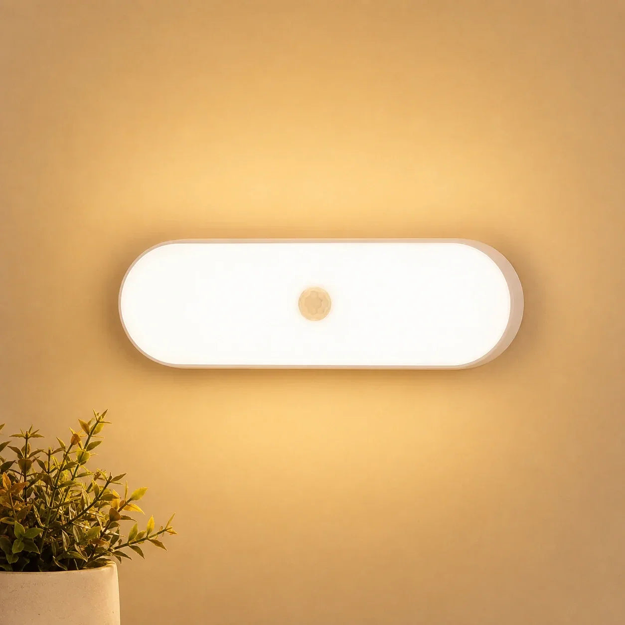 Rechargeable LED wall light with a motion sensor emitting a warm glow, mounted on a wall above a small potted plant in a cosy indoor setting.