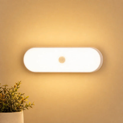 Rechargeable LED wall light with a motion sensor emitting a warm glow, mounted on a wall above a small potted plant in a cosy indoor setting.