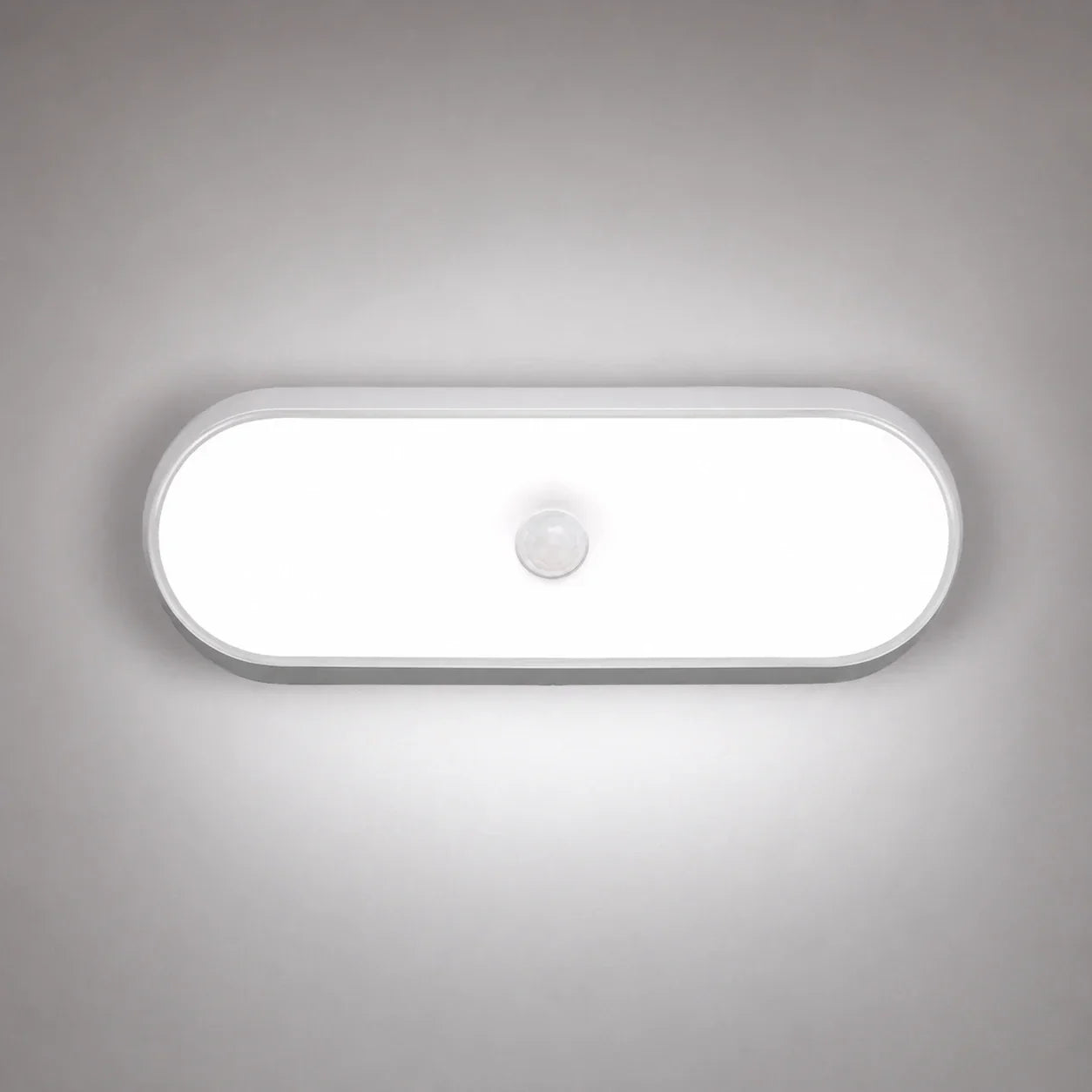 An oval rechargeable motion sensor LED wall light emitting a bright white light, mounted on a grey wall with a central circular sensor.