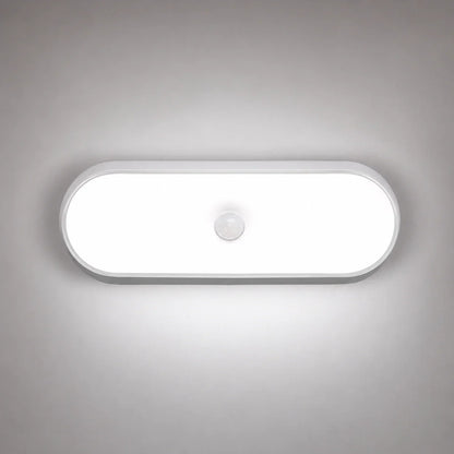 An oval rechargeable motion sensor LED wall light emitting a bright white light, mounted on a grey wall with a central circular sensor.