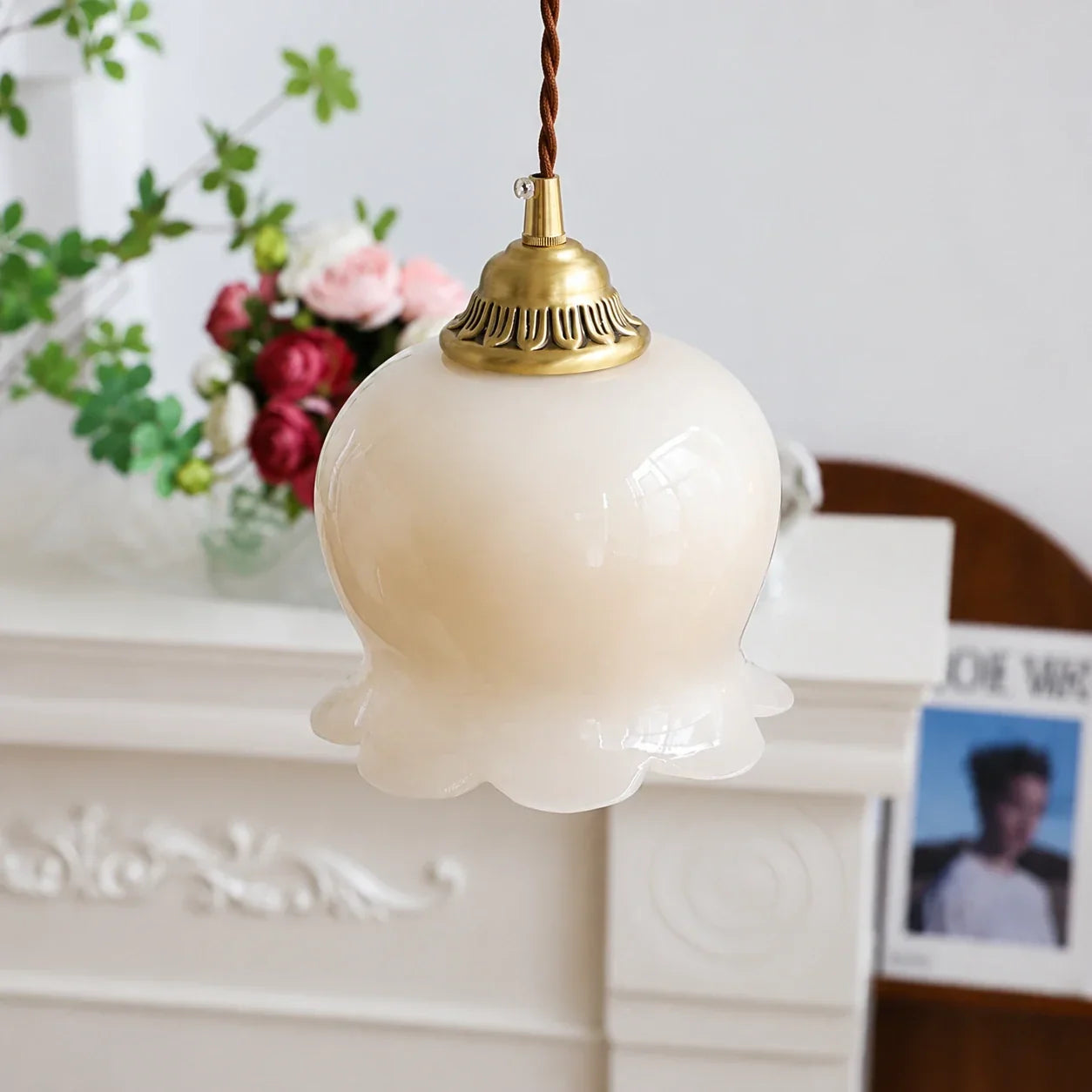 A vintage-style scalloped floral pendant light featuring a cream glass flower-bud shade, brass fixture, and brown braided hanging cord.