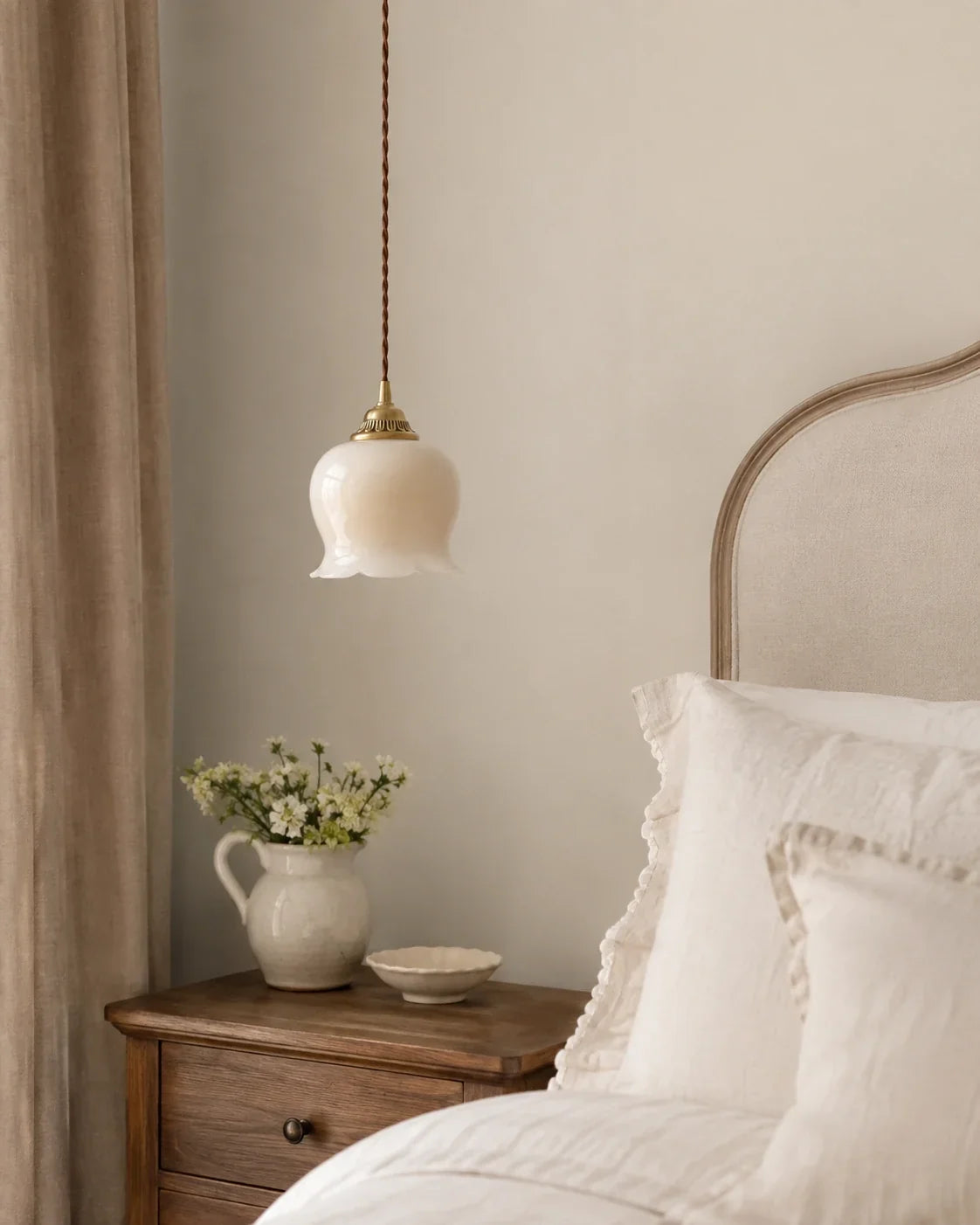 A vintage-style scalloped floral pendant light with a cream glass shade, hanging as bedroom lighting beside a bed with white linen.