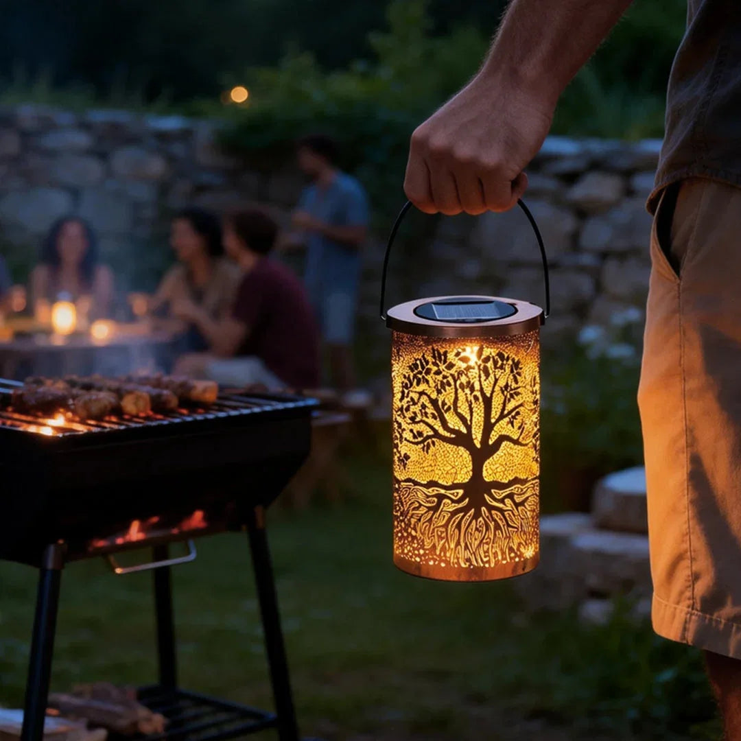 A person holding a glowing copper-coloured Tree of Life solar lantern by its handle, with an outdoor evening barbecue and guests in the background.