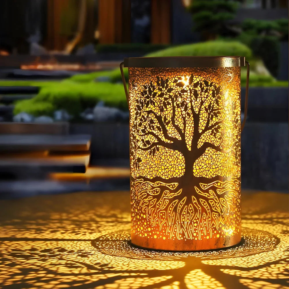 A copper Tree of Life solar lantern glowing with warm light, casting an intricate tree shadow pattern onto a garden surface at night.