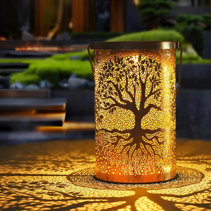 A copper Tree of Life solar lantern glowing with warm light, casting an intricate tree shadow pattern onto a garden surface at night.