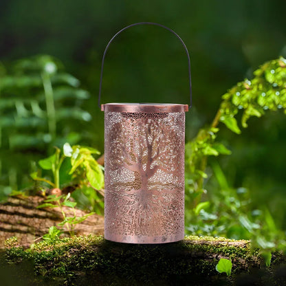 A large copper-coloured Tree of Life solar lantern with a black handle, sitting on a mossy log in a lush green garden background.