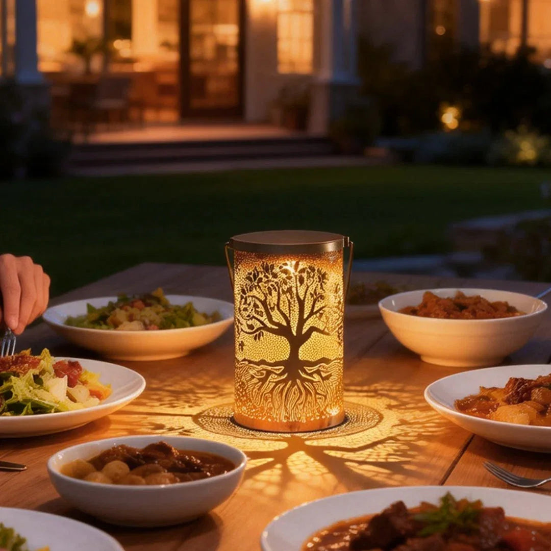 A copper-coloured Tree of Life solar lantern glowing warmly on an outdoor dining table, casting a beautiful tree shadow pattern on the wood.
