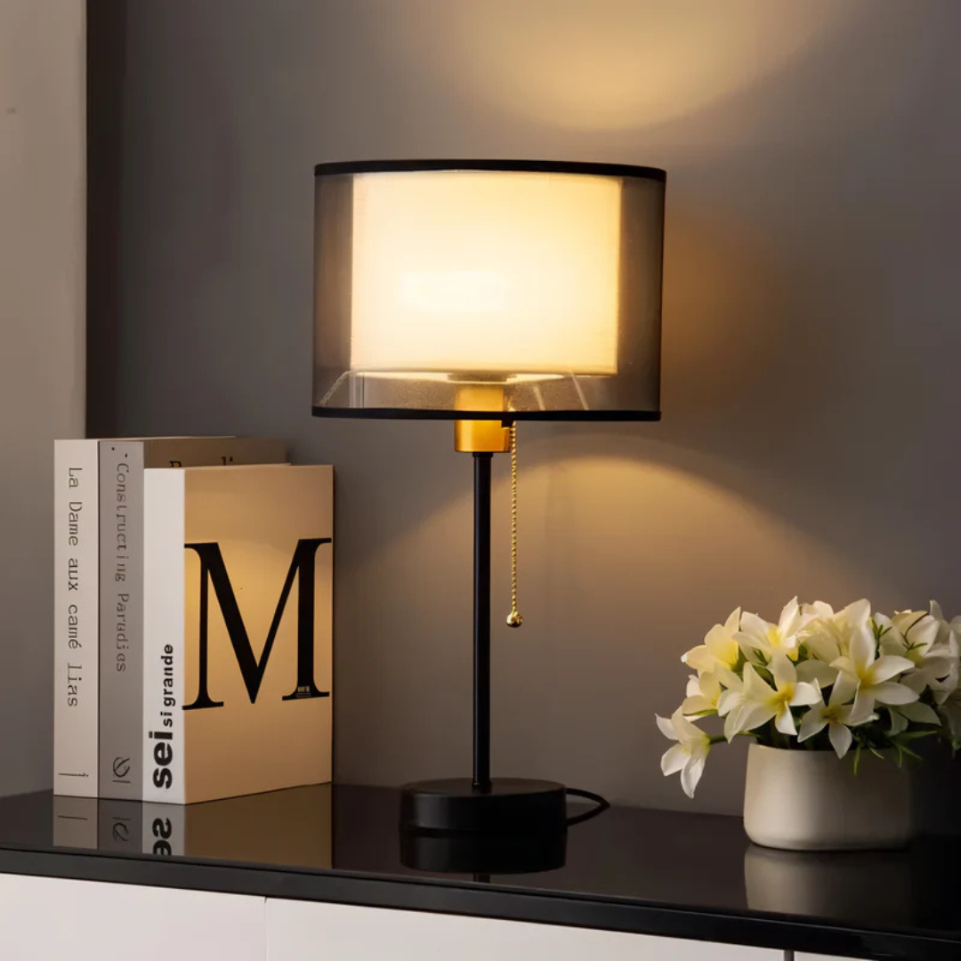 Black vintage table lamp with a transparent double fabric shade beside books and flowers